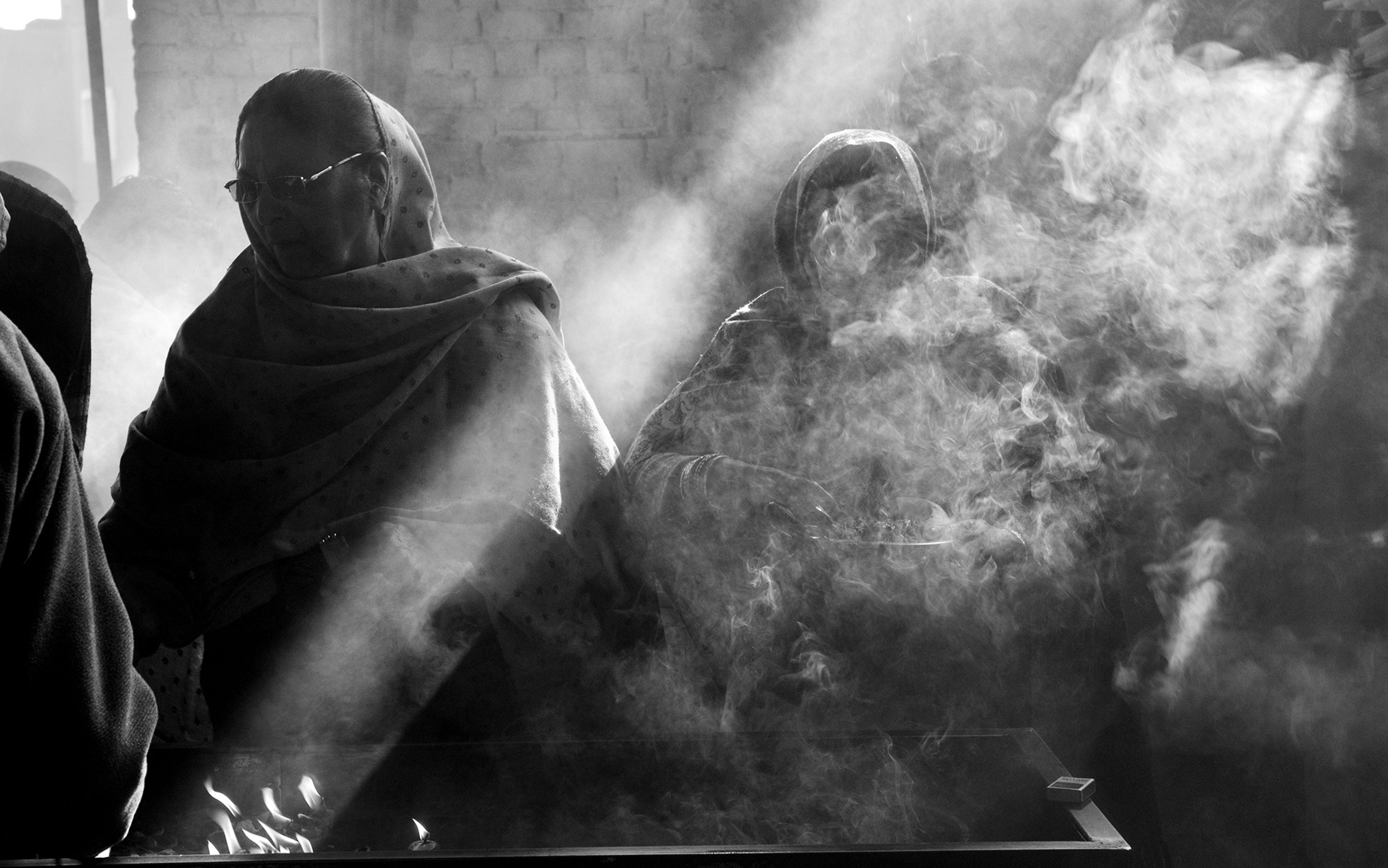 Black and white photo of people wrapped in shawls sitting with light and smoke creating dramatic shadows and highlights.