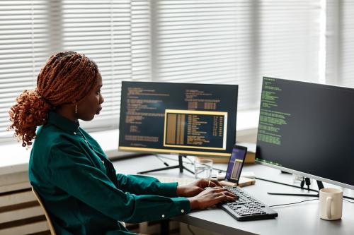 Side view of Black woman IT developer typing on keyboard with programming code on computer screen while working in office