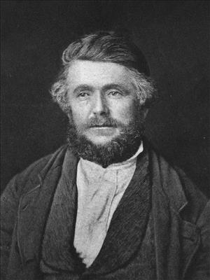 Head and shoulders portrait of a man wearing a dark jacket and scarf, with a mustache, and bushy beard. 